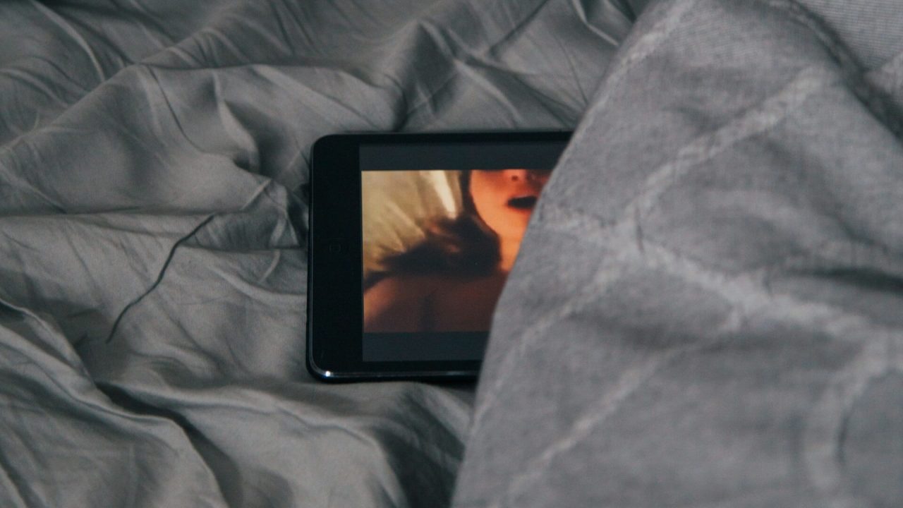 Photo d'un smartphone partiellement placé sous une couverture de lit et dont l'écran affiche une vidéo de divertissement pour adultes.
